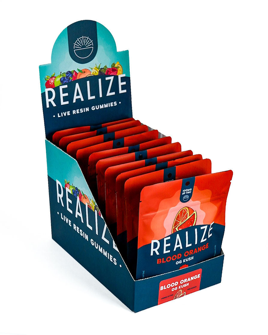Wholesale display box featuring Realize Live Resin Gummies in Blood Orange OG Kush flavor. The display contains bright orange 100mg Δ9 THC gummy pouches with a blood orange illustration and Realize branding, presented in a navy countertop display with a matching Blood Orange OG Kush product label.
